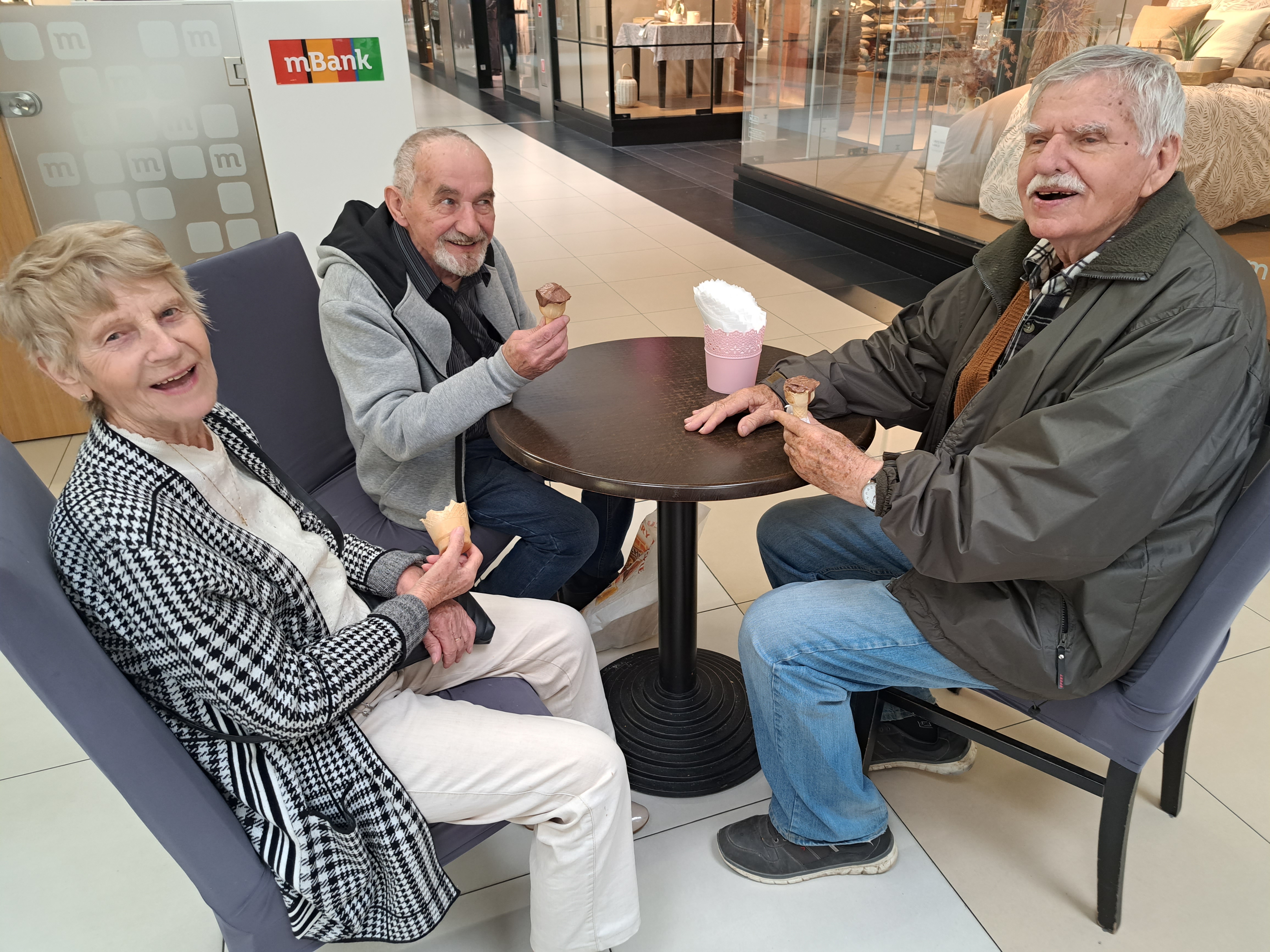 Trójka Seniorów siedzi przy małym stoliku w galerii, jedzą lody